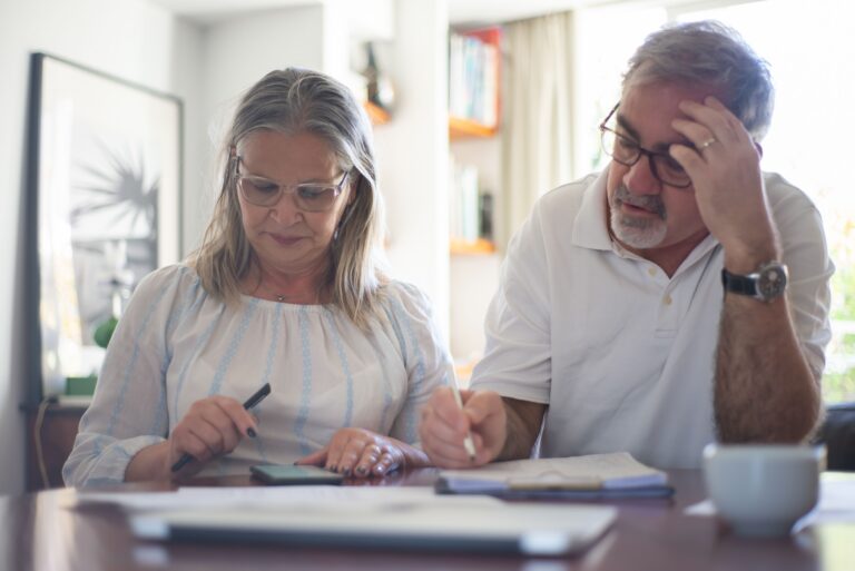 Couple de plus de 50 ans réfléchissant avant une décision financière avec contrat, téléphone, calculatrice et carnet de budget